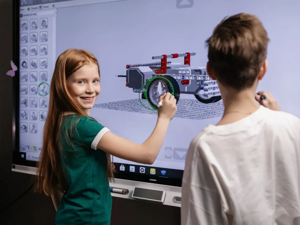 A smiling girl and a boy designing a 3D model on a classroom interactive screen