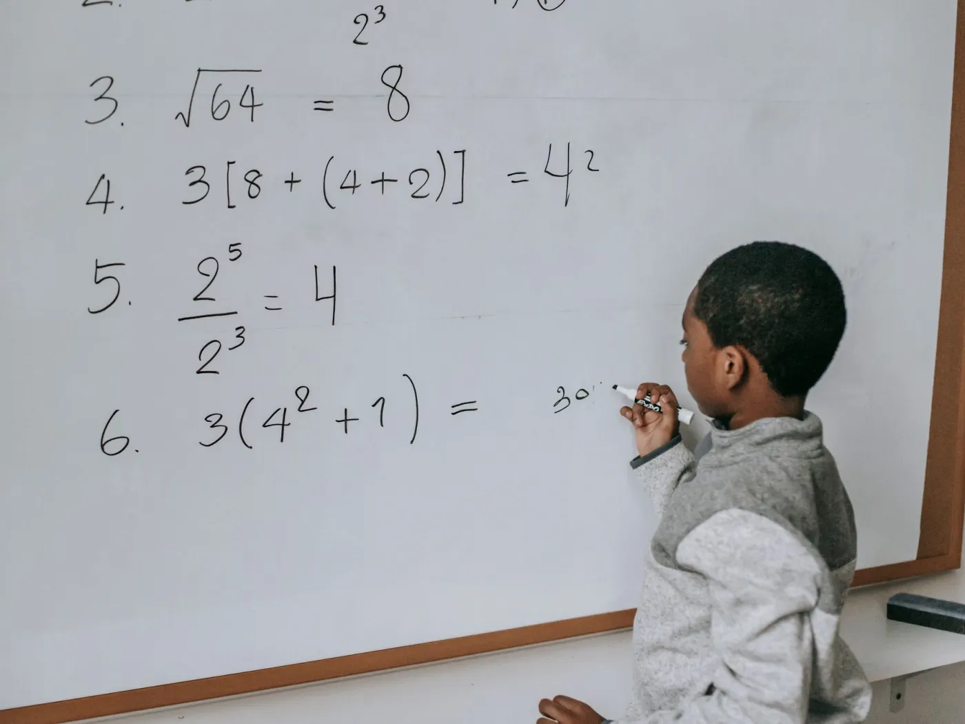 A boy working through exponent and square-root problems with a marker on a classroom whiteboard