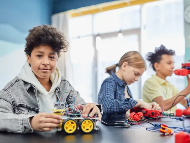 Smiling boy with the robot car he built, classmates working on robotics kits behind him