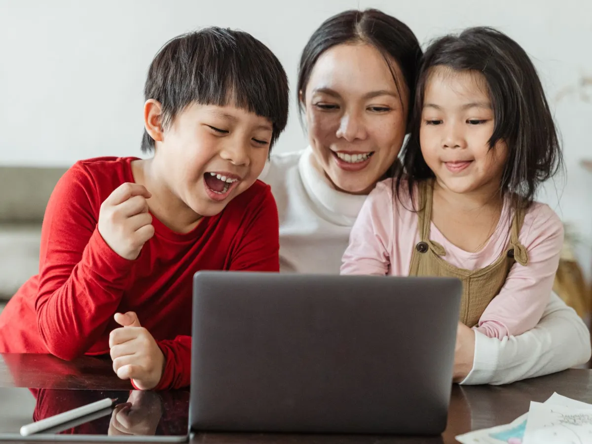 A laughing boy and his little sister cheering at their project on a laptop with their mom