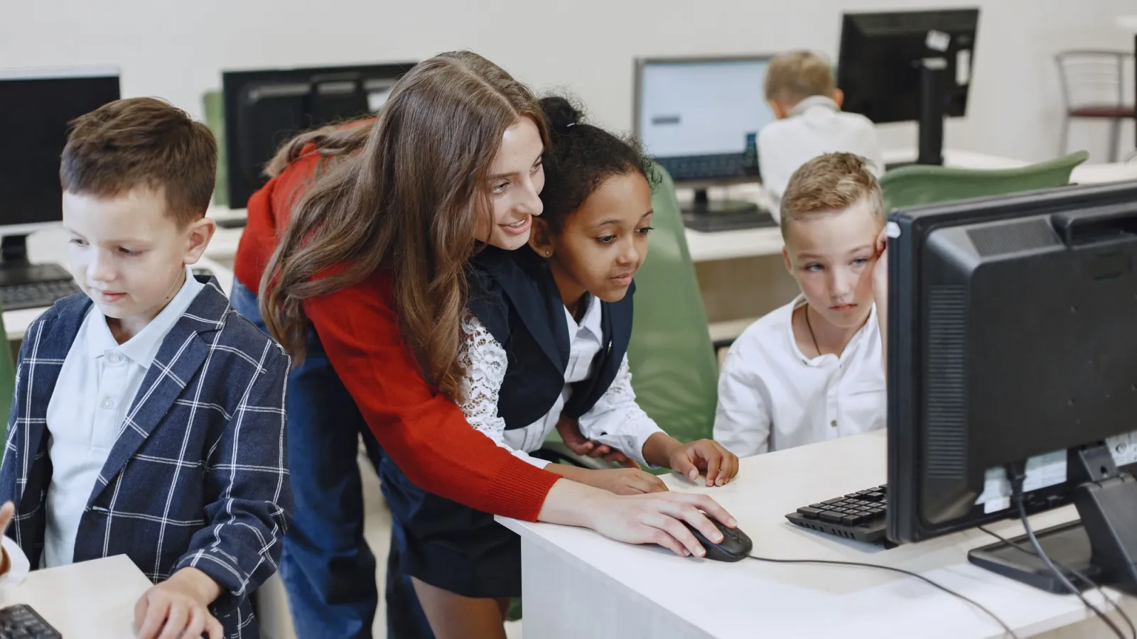 A teacher helping a group of kids with a project at desktop computers in a bright computer classroom